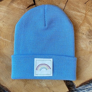 Könnte beinhalten: Eine hellblaue Mütze mit einem kleinen gestickten Regenbogen-Patch auf der Vorderseite.