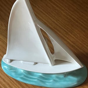 May include: A ceramic sailboat figurine with a white sail and a turquoise base depicting waves. The boat's hull is white, and the mast is a thin, curved line. The figurine is set on a wooden surface.