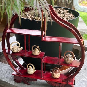 May include: A dark red, wooden display shelf with multiple levels, holding several small, decorative teapots. The shelf has a unique, curved design and is placed in front of a potted plant. The teapots are various colors and styles.