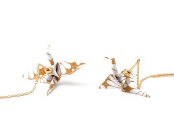 May include: A pair of drop earrings featuring small, folded paper crane charms in white and gold. Each crane is attached to a delicate gold-tone metal chain that extends from the earring post, creating a dangling effect with a metallic finish.