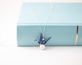 Può includere: Una collana con catena d'argento con un ciondolo a forma di gru origami blu e una perla bianca. Il ciondolo è su un libro blu.
