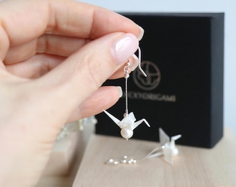 Könnte beinhalten: Zarte Origami-Kranich-Ohrringe mit Perlenakzent. Die Ohrringe hängen an einem silbernen Haken und einer Kette. Eine schwarze Schachtel mit goldfarbenen Buchstaben befindet sich im Hintergrund. Die Ohrringe befinden sich auf einer Holzoberfläche.