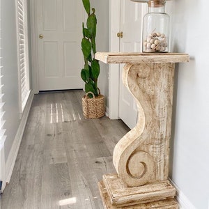May include: Whitewashed wooden console table with a carved scroll design on the leg. The table has a simple rectangular top and is sitting in a hallway with a potted plant and a lamp on top.