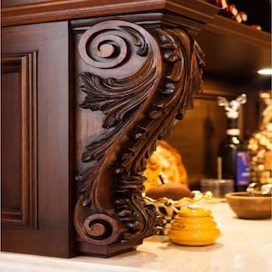 Op de afbeelding: Een donkerbruine houten korbel met ingewikkelde houtsnijwerk van bladeren en spiralen. De korbel is bevestigd aan een kast en is een decoratief element.