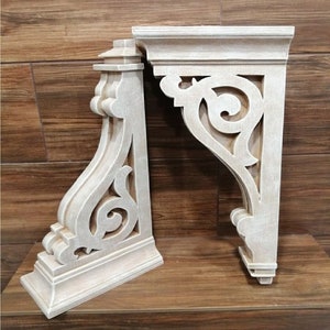 Puede incluir: Dos cornisas de madera sin terminar con intrincados diseños de volutas. Las cornisas son de madera clara y tienen un acabado blanco desgastado.