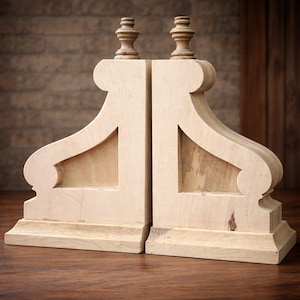 May include: A pair of unfinished wooden bookends. Each bookend features a decorative design with a base, central support, and a curved top with a small, turned finial. The bookends are made of light-coloured wood and are set against a brown background.