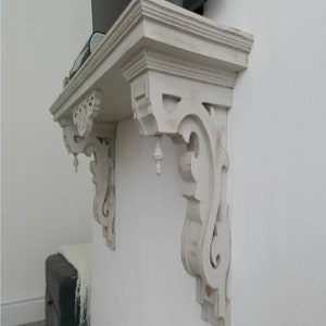 Puede incluir: Soporte de estante de madera pintado de blanco con intrincados adornos y un remate decorativo. El soporte está montado en una pared blanca.