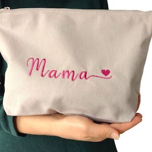 Può includere: Una pochette con cerniera di colore grigio chiaro con la parola "Mama" ricamata in corsivo rosa con un piccolo cuore rosa.