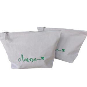 Op de afbeelding: Twee off-white canvas ritszakjes. Op een zakje staat de naam "Anne" geborduurd in groen schrift, met een groen hartsymbool. Het andere zakje heeft het woord "love" geborduurd in groen schrift, ook met een groen hartsymbool.