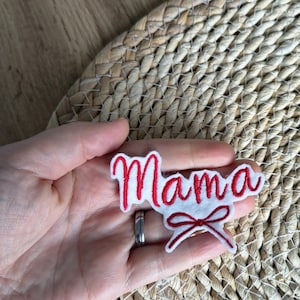 Op de afbeelding: Een wit vilt patch geborduurd met het woord "Mama" in rood cursief schrift. Een rode strik is onder het woord geborduurd. De patch wordt in een hand gehouden, met een geweven achtergrond.
