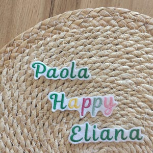 Op de afbeelding: Drie geborduurde naamlabels: "Paola", "Happy" en "Eliana". Elke naam is in witte letters met een groene rand. "Happy" heeft gele en roze accenten. De labels liggen op een geweven, beige oppervlak.