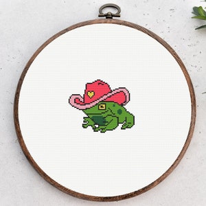 May include: A green frog wearing a pink cowboy hat with a yellow heart on the front. The frog is sitting on a white background.