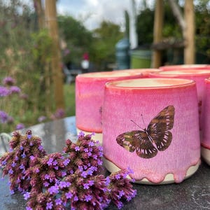 May include: A ceramic mug with a pink glazed exterior featuring a brown butterfly decal. The glaze has a dripping effect near the base, revealing a light-coloured ceramic bottom. The mug is positioned next to a cluster of small purple flowers.
