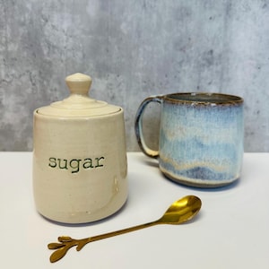 May include: A white ceramic sugar jar with a lid and the word "sugar" written in green on the front. The jar is next to a blue and white ceramic mug and a gold spoon.