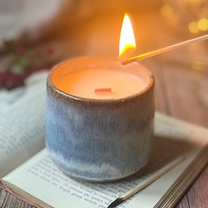May include: A lit candle in a blue and brown ceramic jar with a wooden wick. The candle is sitting on a book with a matchstick next to it.