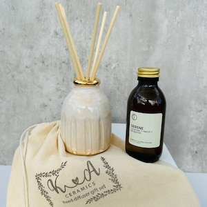 May include: A white ceramic diffuser bottle with gold accents and a set of wooden reeds. The bottle is filled with a clear liquid and is sitting on a beige fabric bag with the text "Kea Ceramics Reed diffuser gift set".