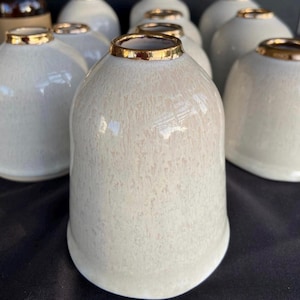May include: A set of six white ceramic cups with a gold rim. The cups have a speckled glaze and a rounded shape.