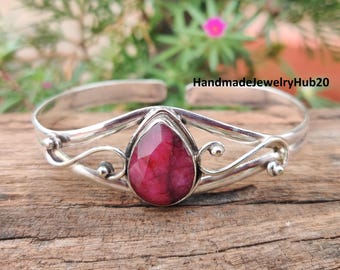 May include: A silver cuff bracelet featuring a large, teardrop-shaped, deep red gemstone. The bracelet has an open design with decorative swirls and small silver beads. The text "HandmadejewelryHub20" is visible.