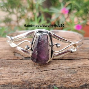 May include: A silver bracelet with a large, rough-cut, purple gemstone at its centre. The bracelet has decorative swirls and small, round accents. The text "HandmadeJewelryHub20" is visible in the background.
