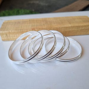 May include: Five silver bangles stacked together. The bangles are smooth and shiny, with a slightly rounded edge.