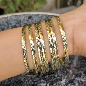 Può includere: Una collezione di braccialetti color oro con una texture martellata. I braccialetti sono impilati su un polso, mostrando la loro superficie riflettente. I gioielli hanno un tono metallico caldo, adatto per l'uso quotidiano o occasioni speciali.