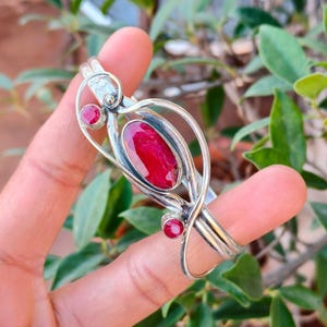Peut inclure: Un bracelet argenté orné de trois pierres précieuses rouges vives. Le bracelet présente un design ouvert avec une pierre centrale ovale et deux petites pierres rondes. Le bracelet est présenté sur un fond de feuillage vert.