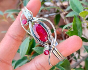 May include: A silver bracelet with three vivid red gemstones. The bracelet has an open design with an oval central stone and two smaller round stones. The bracelet is displayed against a backdrop of green leaves.