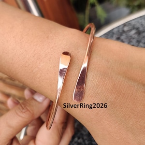 May include: A rose gold coloured bangle bracelet with a simple, curved design. The bracelet is open and adjustable to fit different wrist sizes.  SilverRing2026 is printed on the bracelet.