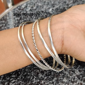 Puede incluir: Un conjunto de cinco pulseras de plata, incluyendo una pulsera trenzada, en la muñeca de una persona.