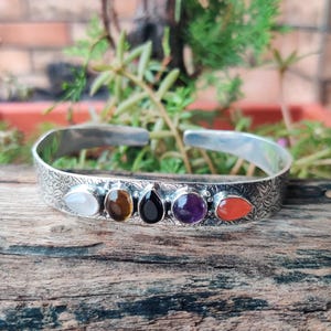 Könnte beinhalten: Silbernes Armreif-Armband mit eingraviertem Blumenmuster. Das Armband hat sechs farbige Edelsteine: weiß, braun, schwarz, lila und orange. Das Armband wird auf einer Holzoberfläche präsentiert.