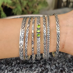 May include: A set of six silver bangles with intricate designs. The bangles are stacked on a person's wrist and feature green gemstones.