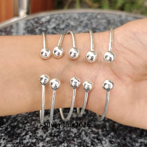 May include: Six silver bangle bracelets with round ball ends. The bracelets are stacked on a person's wrist.