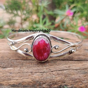 May include: A silver bracelet with an oval, faceted, deep red gemstone at its center. The bracelet has decorative swirls and small silver beads. The text "HandmadeJewelryHub20" is visible.