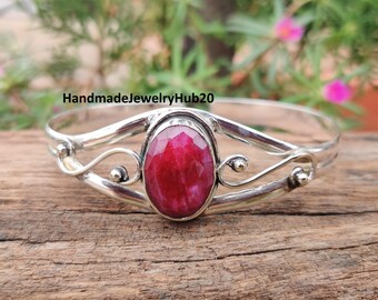 May include: A silver bracelet featuring a large, oval-cut, deep red gemstone. The bracelet has decorative swirls and small silver beads. The text "HandmadeJewelryHub20" is visible.