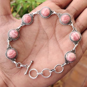 Peut inclure: Un bracelet en argent orné de huit pierres roses rondes. Chaque pierre est sertie dans une lunette décorative en forme de corde. Le bracelet est doté d'un fermoir à bascule et est présenté sur un fond neutre.