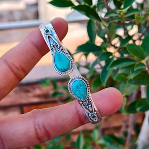Peut inclure: Un bracelet manchette en argent avec deux pierres turquoise en forme de goutte d'eau. Le bracelet a un design ouvert avec des détails ornés autour des pierres et le long du bracelet. Les pierres turquoise ont un aspect moucheté. Le bracelet est tenu par une main.