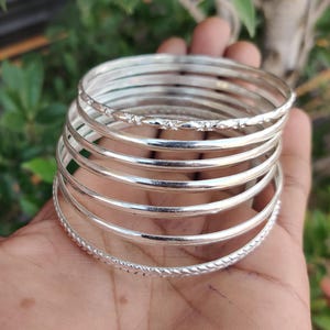 May include: A stack of silver bangles, some with textured designs, are held in a hand. The bangles are round and shiny, reflecting light. The background is blurred green foliage.