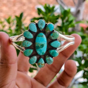 May include: A silver cuff bracelet featuring a large central oval turquoise stone surrounded by twelve smaller pear-shaped turquoise stones. The stones display natural veining in shades of blue and green, set within a polished silver frame.