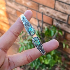 Puede incluir: Brazalete de puño plateado con dos secciones en forma de lágrima. Cada sección presenta una incrustación de concha de abulón iridiscente con tonos azules, verdes y turquesas. El brazalete tiene detalles ornamentados y un borde tipo cuerda.