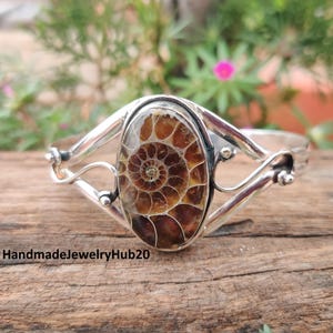May include: Silver cuff bracelet with an oval ammonite fossil. The fossil has a detailed spiral pattern in brown and tan. The bracelet is set against a natural wood surface. The text "HandmadeJewelryHub20" is visible.