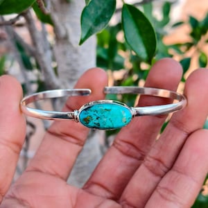 Könnte beinhalten: Ein silbernes Armreif-Armband mit einem leuchtenden Türkisstein in der Mitte. Der oval geformte Stein hat eine leuchtende türkisfarbene Farbe mit braunen und schwarzen Einschlüssen. Das Armband wird in einer Hand vor einem Hintergrund aus grünen Blättern gehalten.