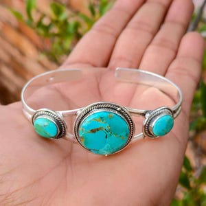 May include: Silver cuff bracelet with three turquoise stones. The bracelet has a polished silver finish, with a large central turquoise stone and two smaller stones. The turquoise stones have a mottled blue-green appearance.