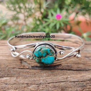 May include: A silver-toned bracelet with a large, round turquoise stone at the center. The stone is a vibrant blue-green color with a textured surface. The bracelet has a delicate, open design with swirling details.