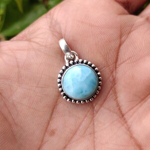 May include: A round, light blue larimar pendant with a silver-colored bail. The gemstone is set in a silver-colored bezel with a beaded border. The pendant is held in a hand, showcasing the stone's color and design.