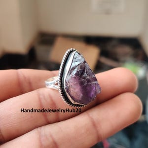 Könnte beinhalten: Ein Silberring mit einem großen, rohen Amethyst-Edelstein. Der Amethyst hat eine tiefviolette Farbe mit einer strukturierten Oberfläche. Der Ring hat ein gedrehtes Seil-Design um den Stein und den Text "HandmadeJewelryHub20".