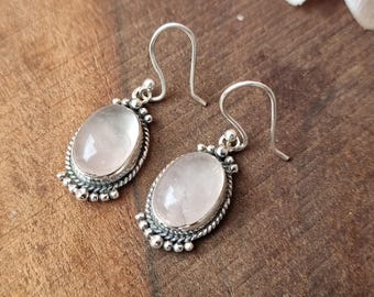 May include: A pair of silver earrings with oval, light pink gemstones. The gemstones are set in a silver frame with a twisted rope design and small silver beads around the edge. The earrings have a hook closure.