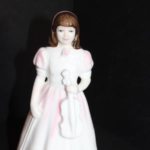 May include: A porcelain figurine of a girl in a white dress with pink accents, holding a white violin. The figurine has brown hair and a pink headband. A pink rose is on the chest. The figurine is a decorative item.