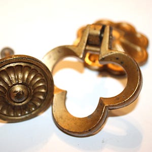 May include: Close-up of two antique brass drawer pulls. One is a round knob with a sunburst design, and the other is a clover-shaped pull. The hardware has a worn, vintage appearance.