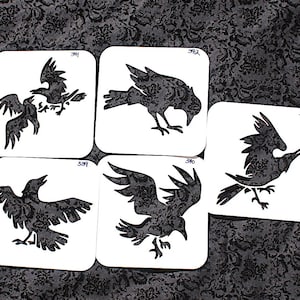 Puede incluir: Cinco posavasos cuadrados blancos con ilustraciones de cuervos negros. Cada posavasos presenta un diseño de cuervo diferente, con variaciones en la pose y la posición de las alas. Los posavasos están dispuestos sobre una tela oscura estampada.