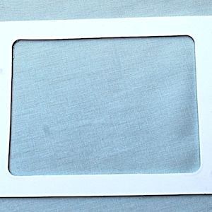 May include: A white rectangular frame with a cutout rectangle in the center. The frame is on a light blue fabric surface. The frame has rounded corners.
